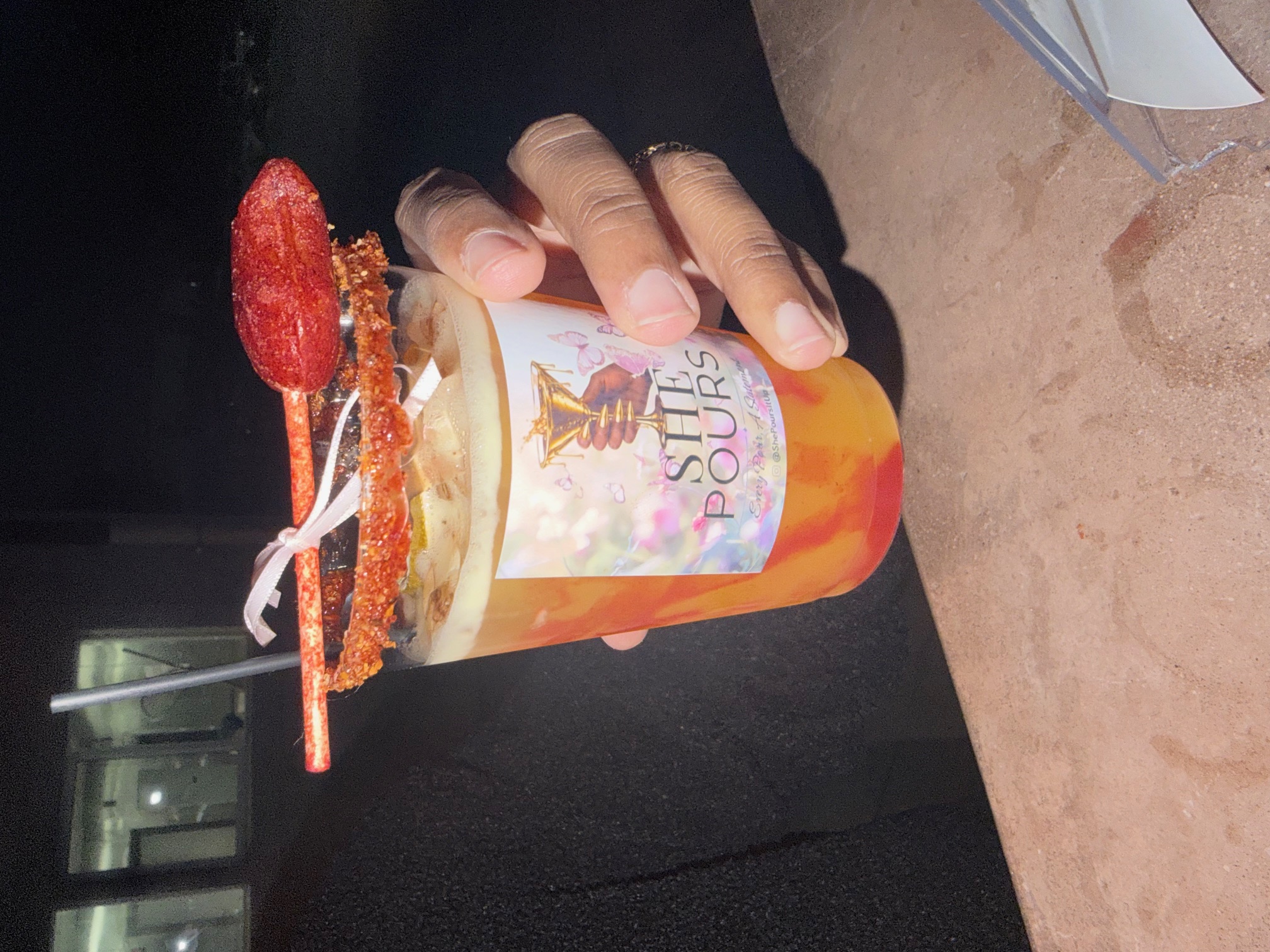 Signature chili-rim cocktail with Lucas candy garnish in a She Pours branded cup, held by Ashlee.